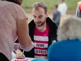 A photograph of Zak Viney at the Hanley Park event. Zak is white with brown hair and a brown beard, wearing a pink Living Streets tshirt. Zak is sat down at a stall and surrounded by other people in the foreground and background who are out of focus.