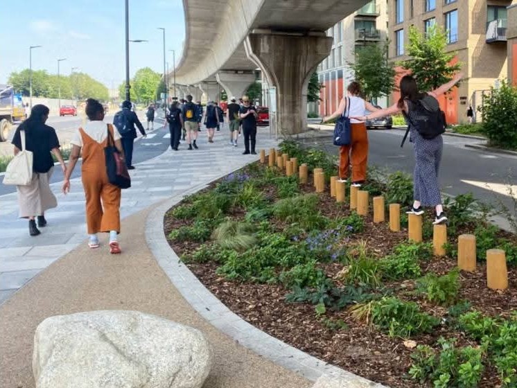 A photograph of a pedestrianised underpass on a clear, sunny day. People are walking on the pavements, and balancing on bollards in a small pocket garden.