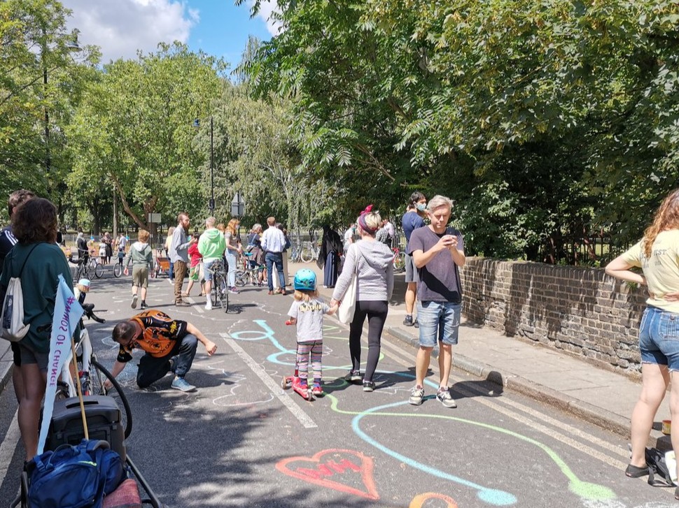 A sunny pedestrianised street filled with adults and children walking. There are trees lining the street either side. The street tarmac is covered in bright and colourful chalked designs.